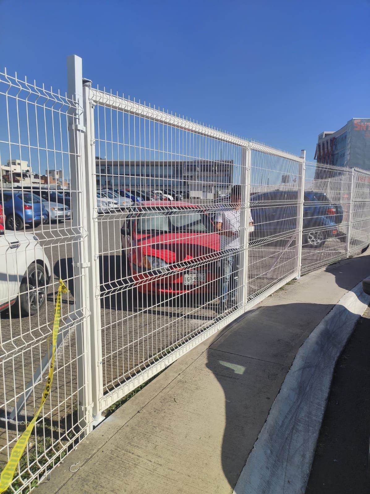 Una valla metálica blanca con coches aparcados visibles al otro lado. Una persona está de pie detrás de la valla.