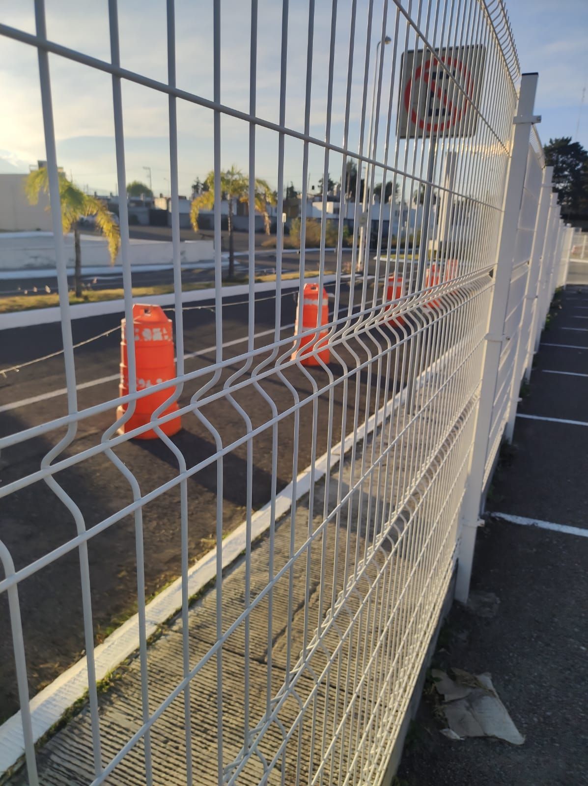 Cerca de alambre blanco a lo largo de un estacionamiento con conos naranjas.