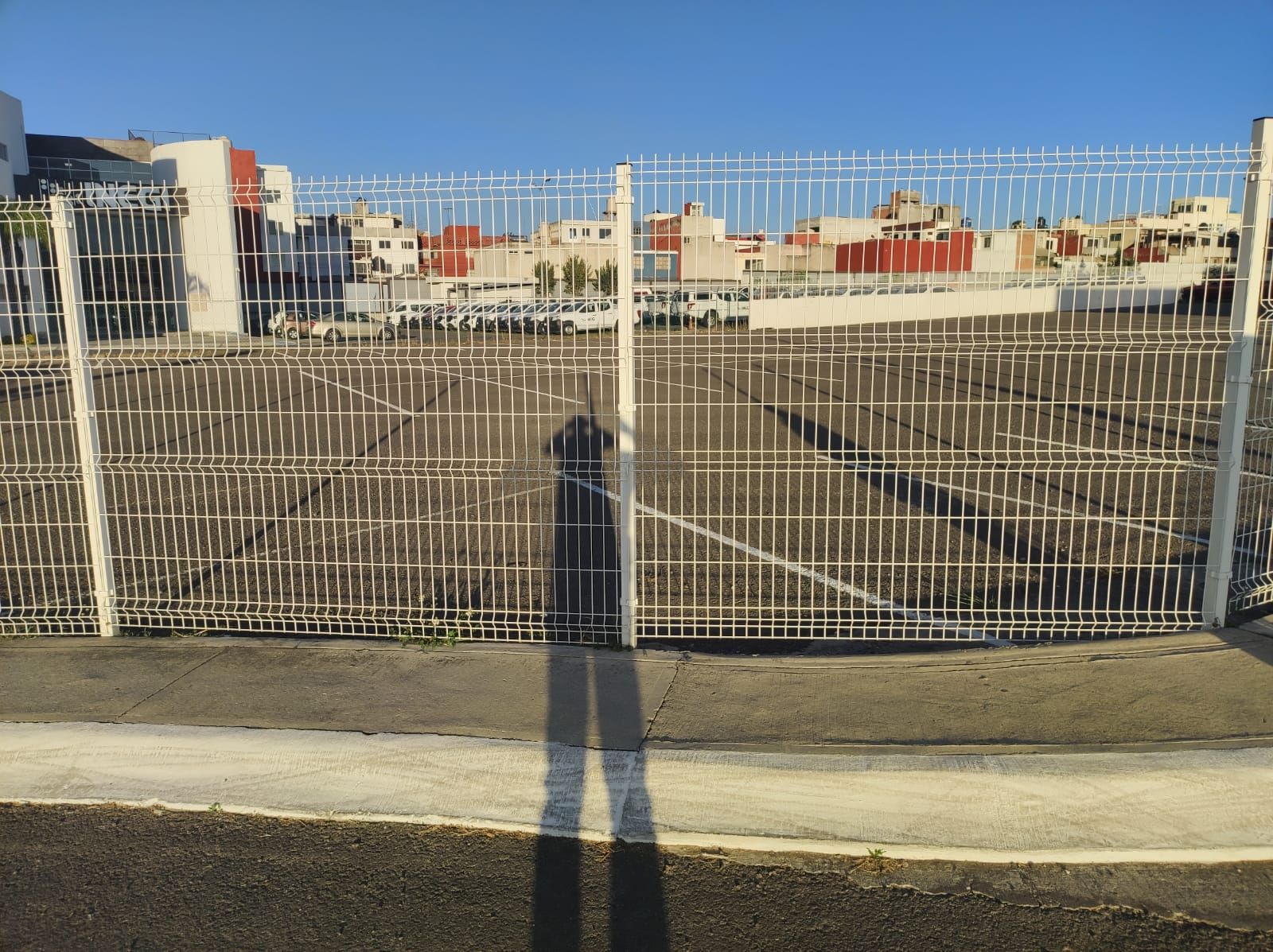 Sombra de persona delante de una valla metálica blanca, edificios al fondo.
