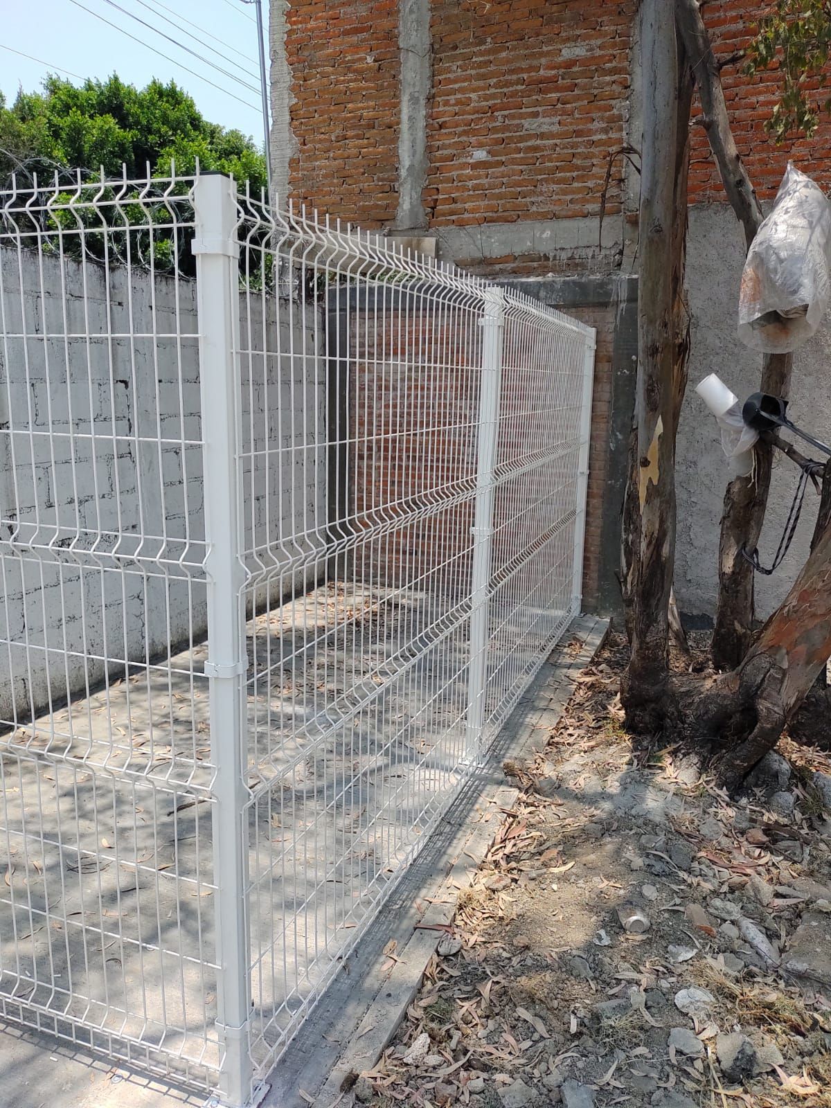 Valla de alambre blanco delante de una pared de ladrillos y un edificio, con un árbol a la derecha.