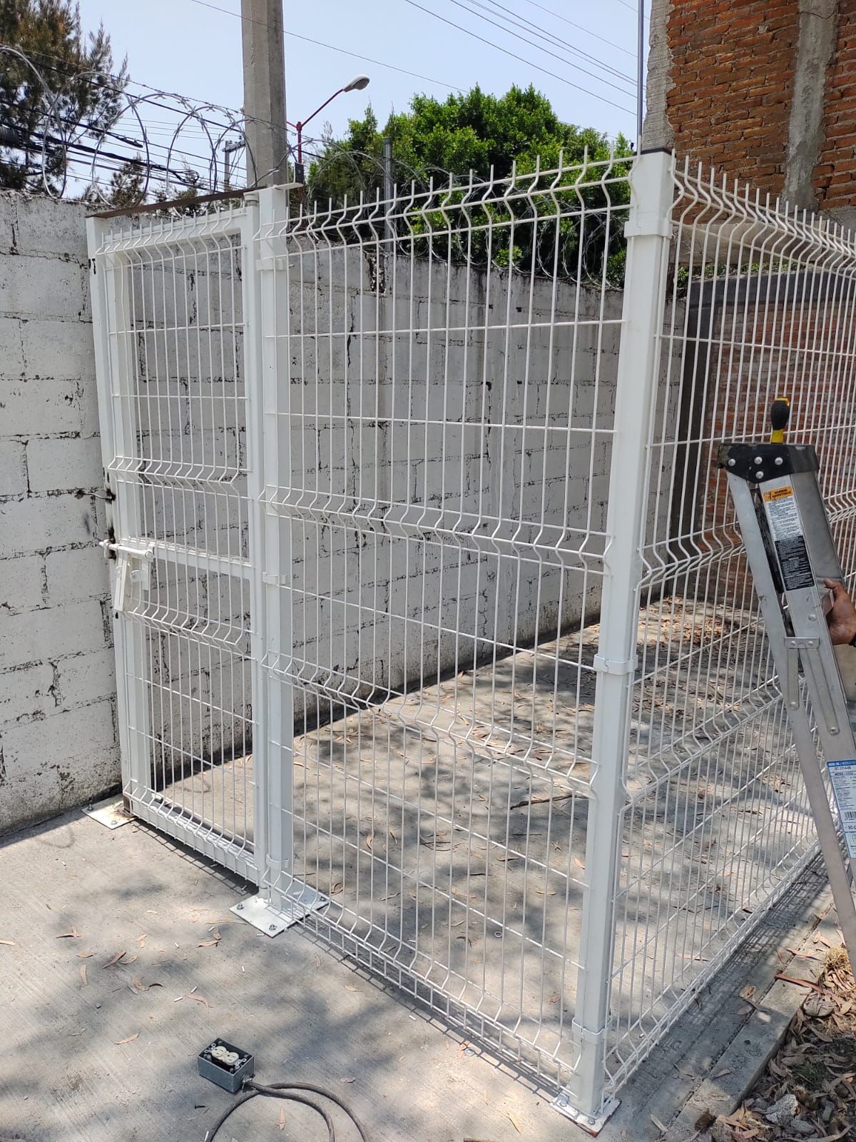 Puerta de seguridad metálica blanca con malla metálica soldada, fijada en hormigón. Hay una escalera junto a la puerta.