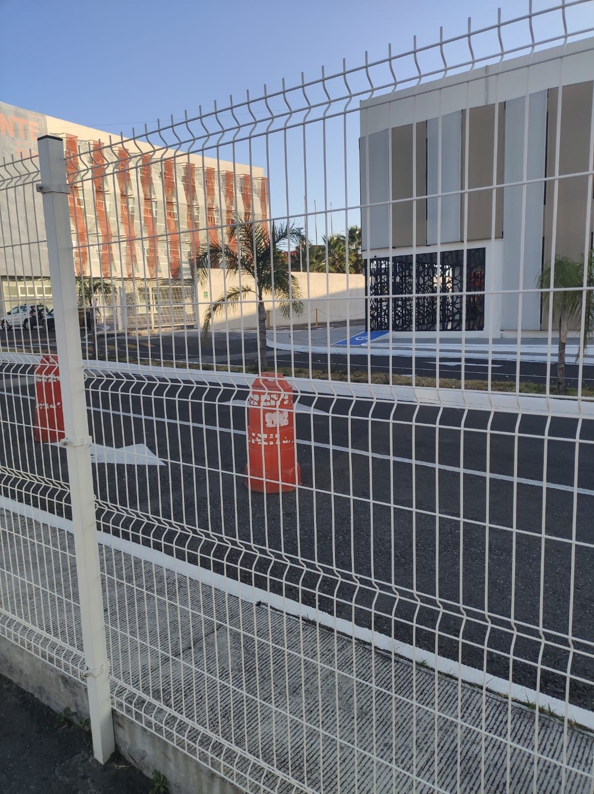 Una valla blanca frente a dos edificios. Conos de tráfico naranjas bordean la carretera asfaltada entre los edificios.