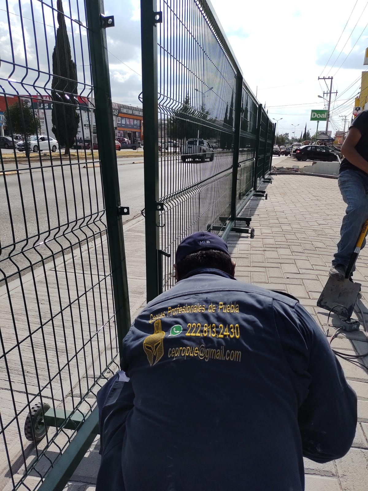 Persona instalando una cerca de alambre verde en una acera, con una calle muy transitada al fondo.