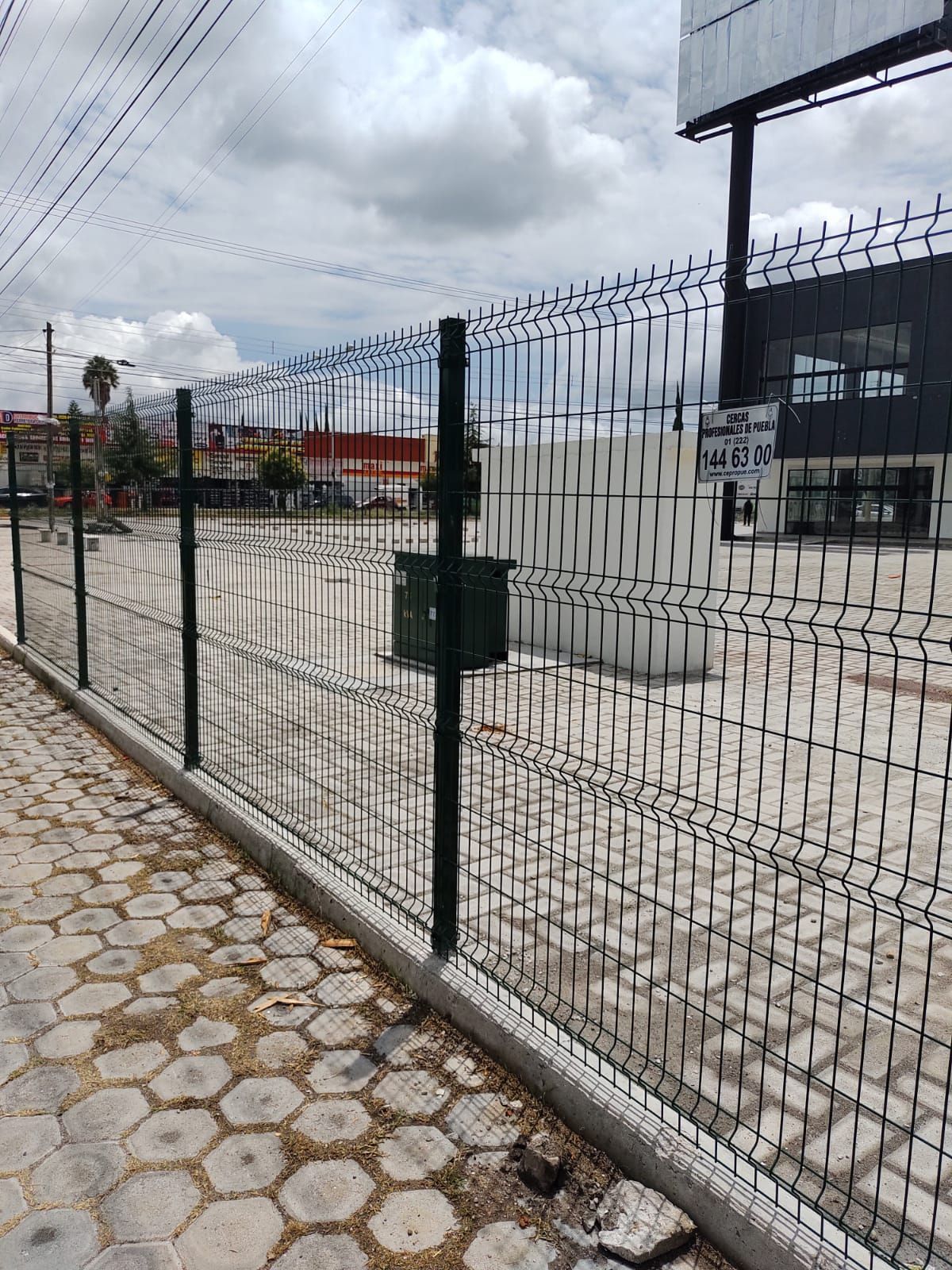 Una cerca de malla metálica rodea un terreno baldío con un buzón en un poste. Cielo nublado.
