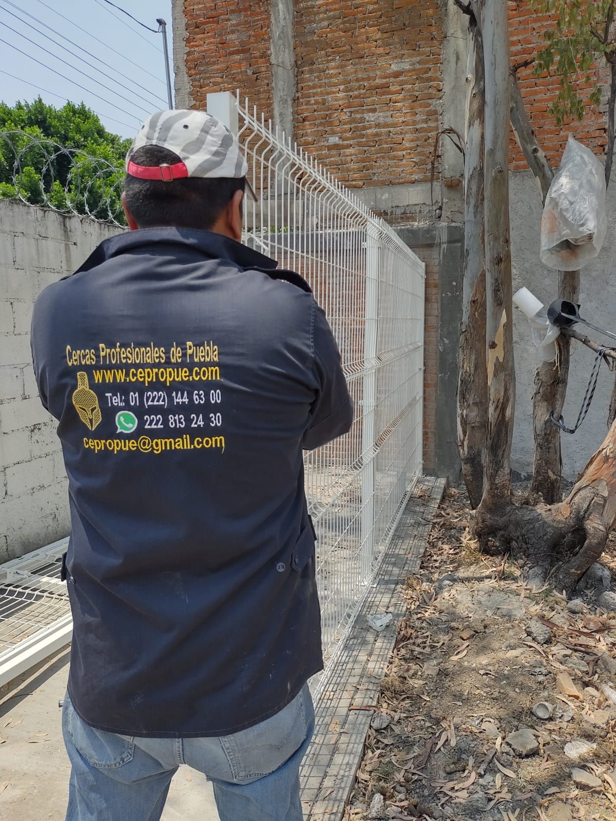 Hombre con uniforme de trabajo mirando una puerta de metal blanca, al aire libre, cerca de una pared de ladrillos.