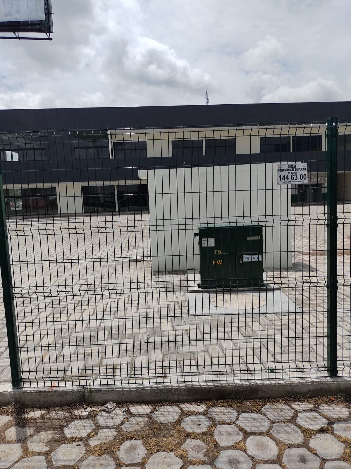 Una caja verde de servicios públicos tras una cerca de alambre. Un edificio con ventanas oscuras al fondo. Cielo nublado.