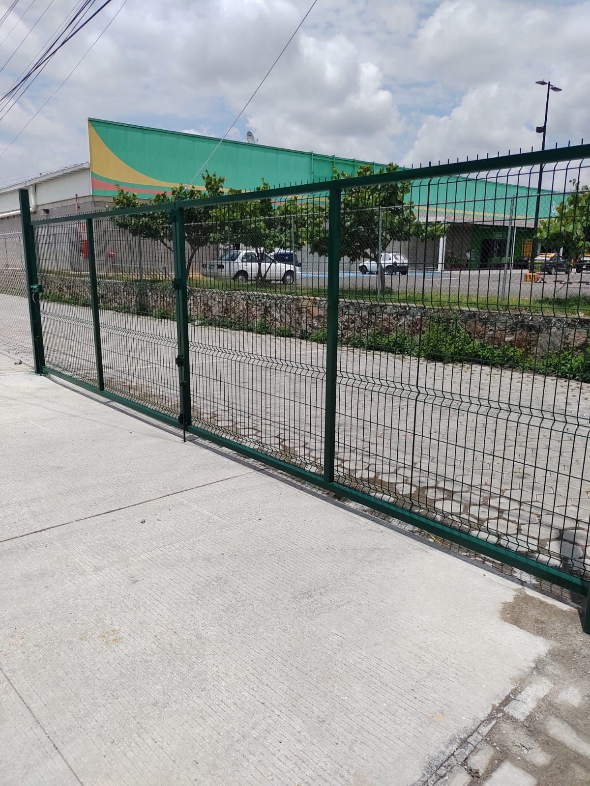 Valla metálica verde sobre una acera de hormigón, frente a un edificio con árboles y vehículos en un día soleado.