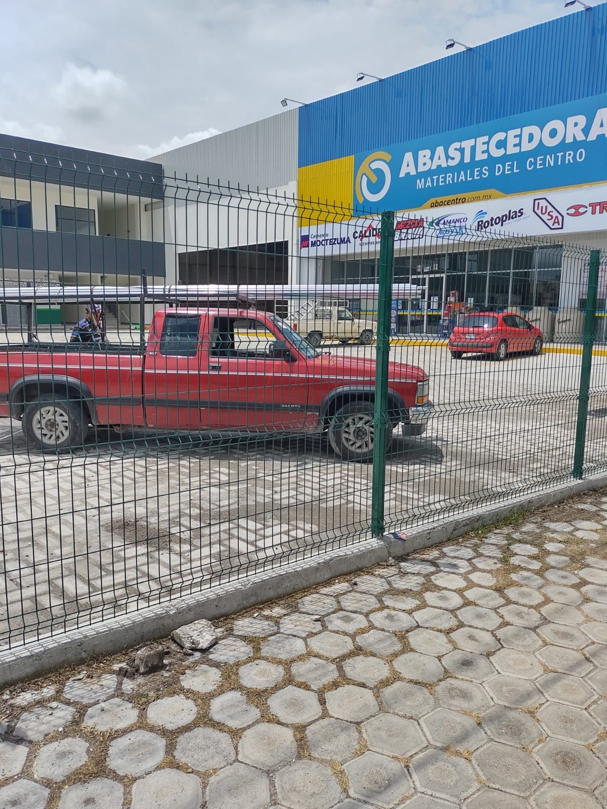 Camioneta roja estacionada frente a una tienda, Abastecedora, con letrero azul. Cerca metálica y acera hexagonal.