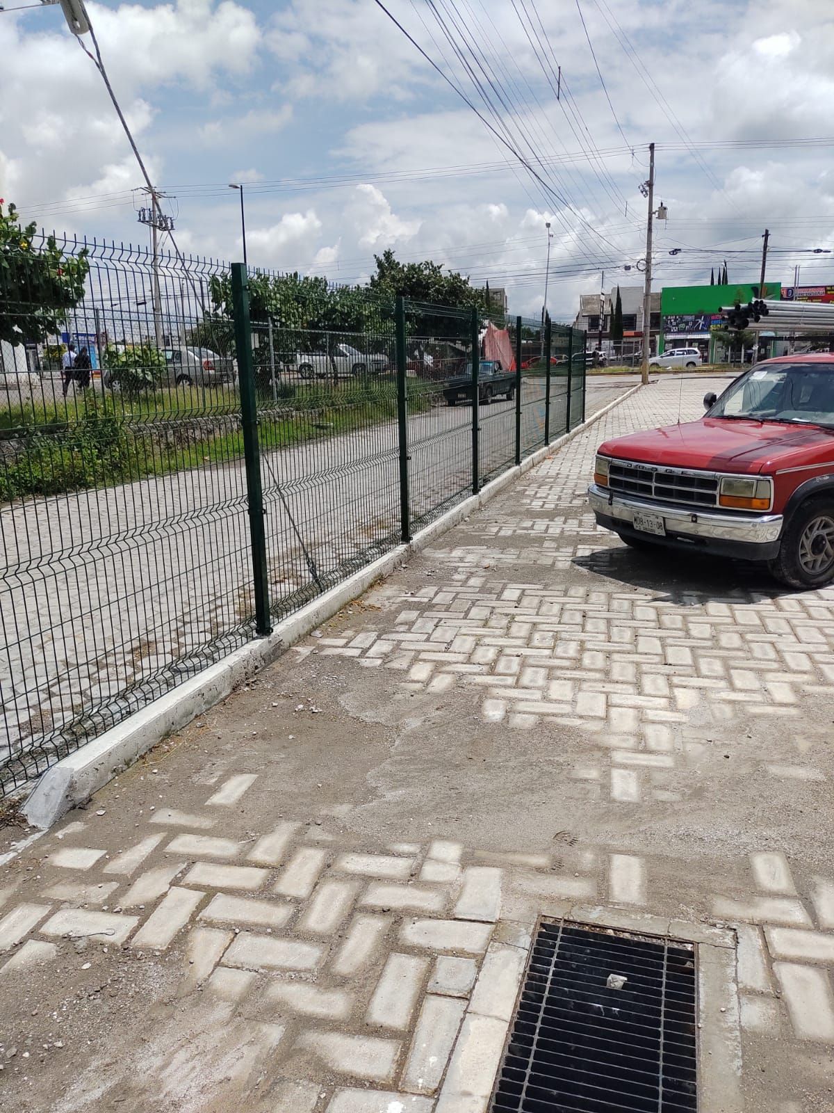Camión rojo estacionado junto a una cerca de alambre a lo largo de una pasarela pavimentada con ladrillos; entorno urbano.