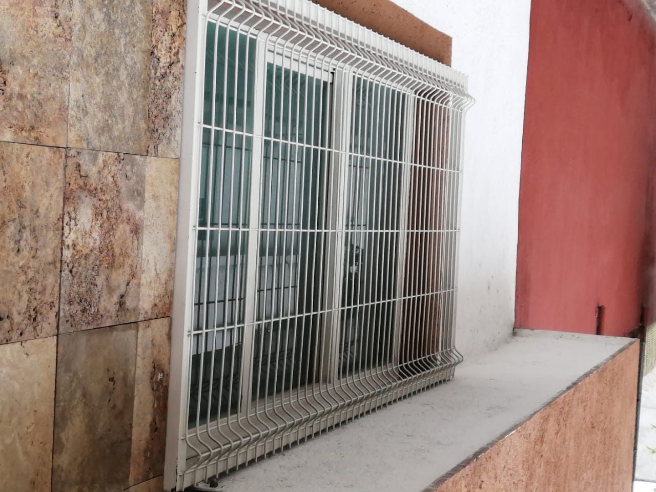 Ventana con rejas metálicas de seguridad, empotrada en pared de piedra y color blanco.