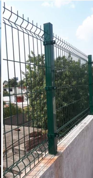 Valla de alambre verde sobre una cornisa de hormigón, con un poste cuadrado. El fondo incluye árboles y edificios.