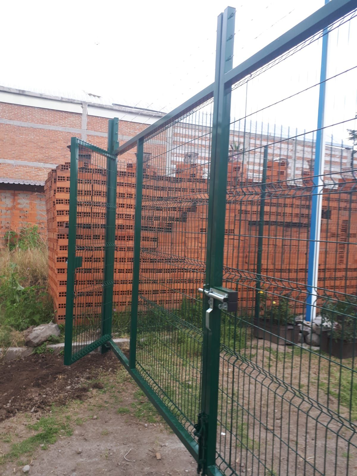 Puerta metálica verde abierta, con pared de ladrillo y valla al fondo.