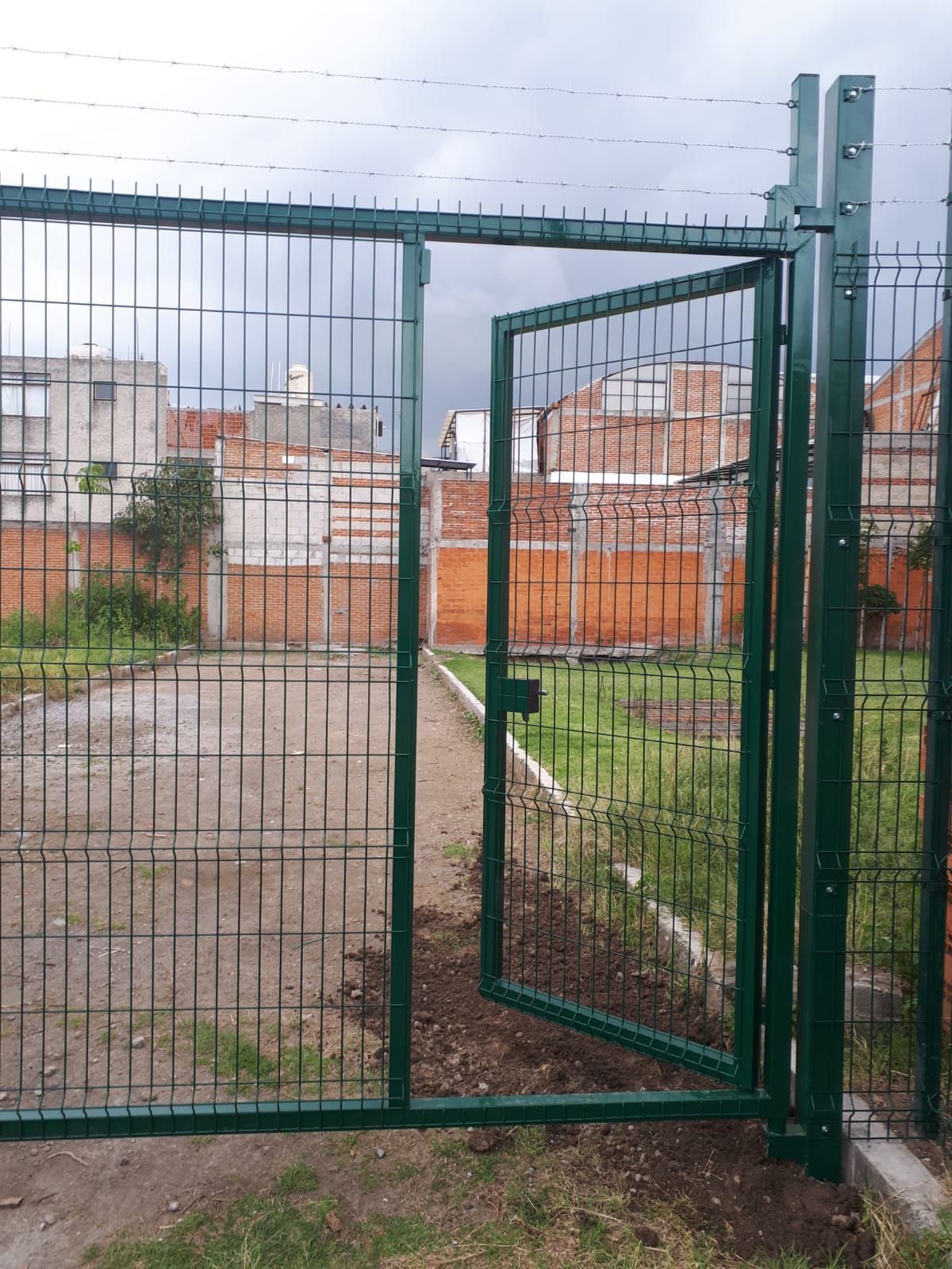 Puerta metálica verde, parcialmente abierta, que conduce a una zona verde con casas al fondo.