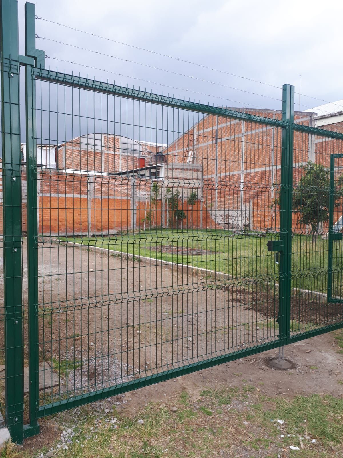 Puerta metálica verde con rejas verticales, abierta, que conduce a un terreno con césped y edificios de ladrillo.