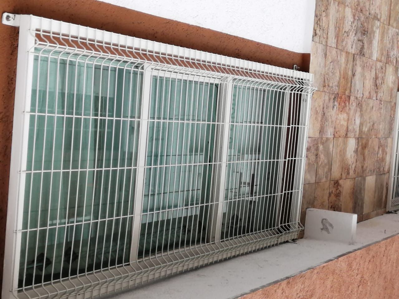 Ventana con rejas de seguridad metálicas blancas. La ventana tiene vidrio y se encuentra en un edificio de ladrillo y paredes pintadas.