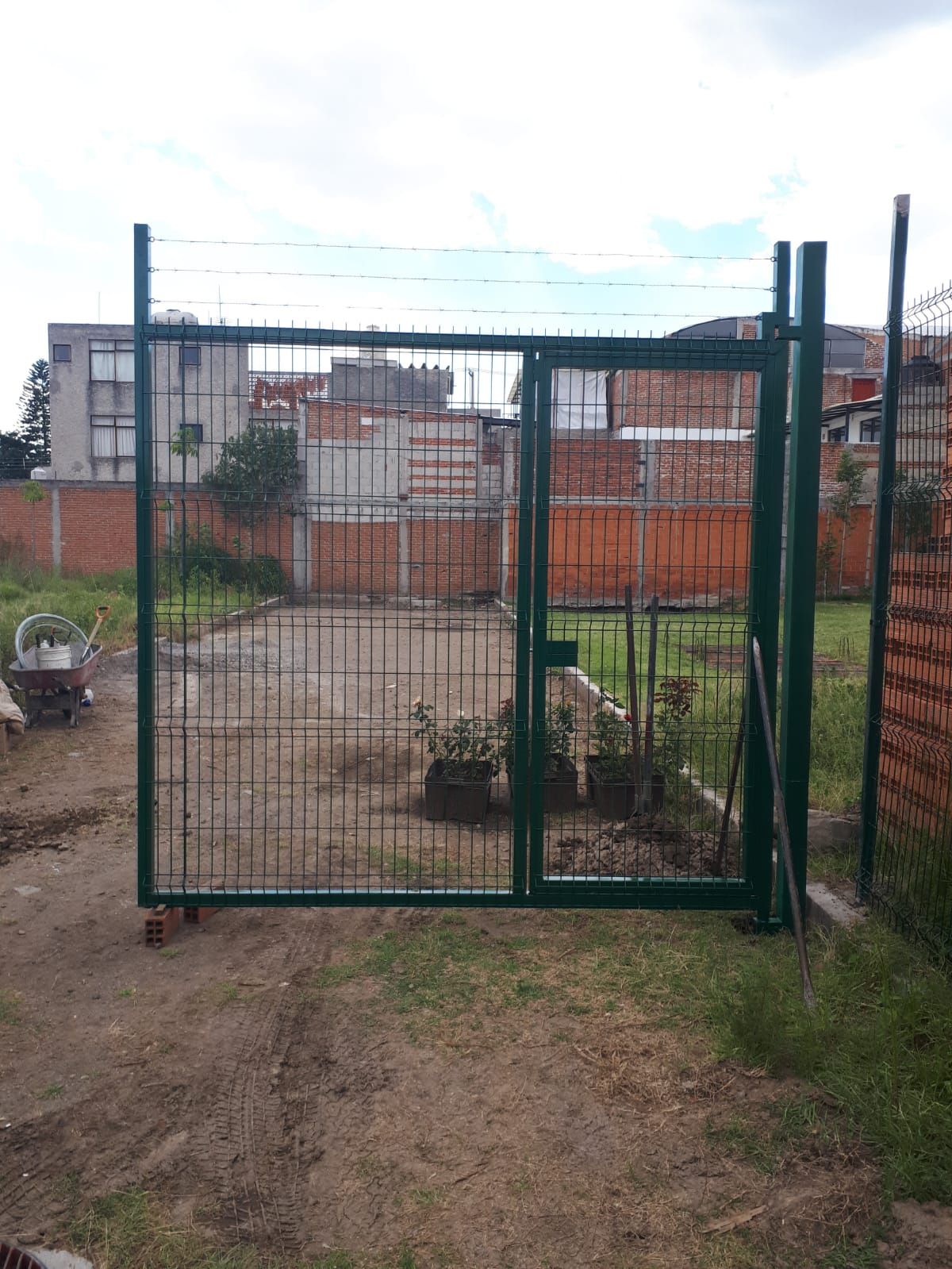 Puerta de metal verde con cerco de malla de alambre frente a un patio de tierra y edificios.