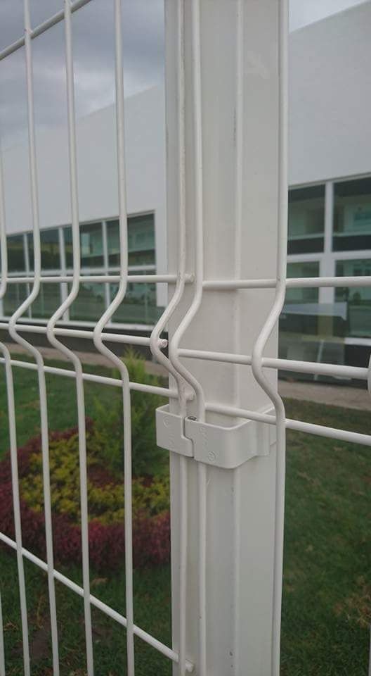 Valla metálica blanca con postes cuadrados, conectados por alambres horizontales, frente a un edificio y césped.