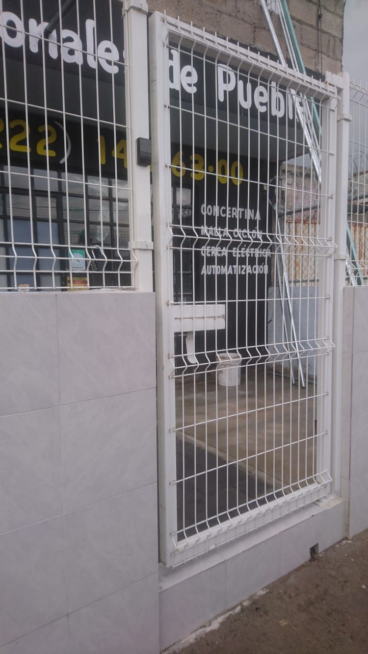 Puerta de seguridad de metal blanco frente a un edificio; texto visible detrás de ella.