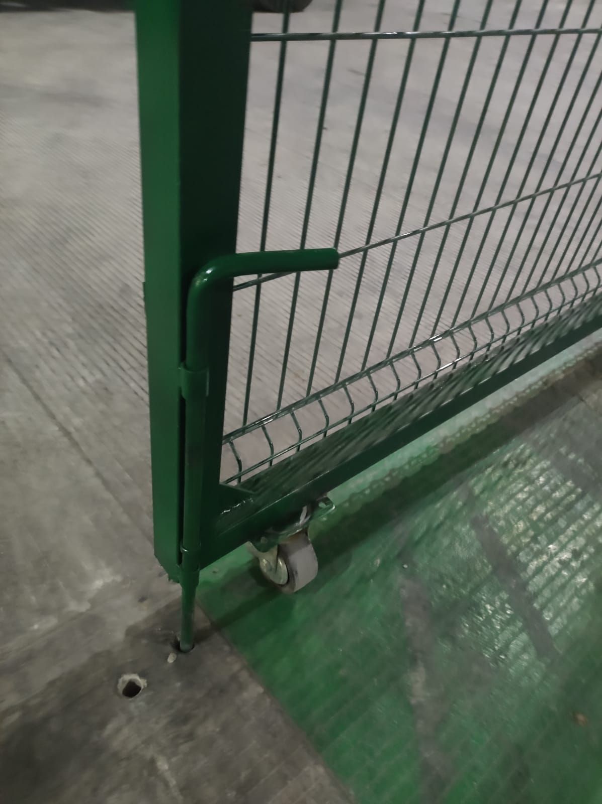 Puerta metálica de color verde con malla de alambre y manija, colocada sobre un piso de concreto con rueda y pasador.