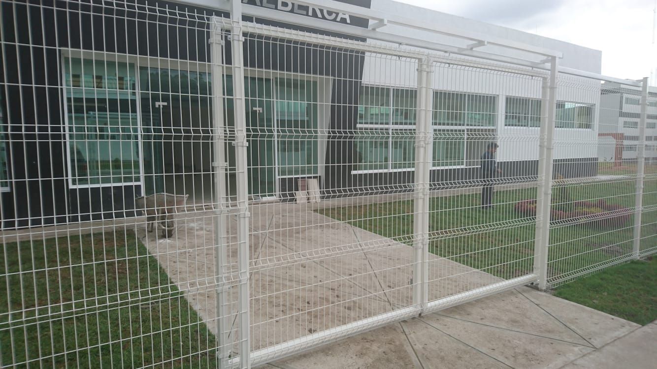 Puerta con barrotes blancos frente a un edificio moderno con grandes ventanales. Zona de césped en primer plano.