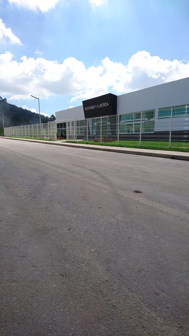 Vista exterior del edificio “ALFA UNIS”, un edificio industrial de color claro con un cartel negro, una calle y una colina verde.