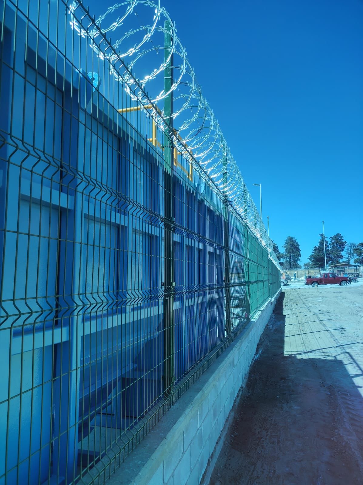 Una valla de seguridad coronada con alambre de púas, que protege un edificio azul contra un cielo azul claro.