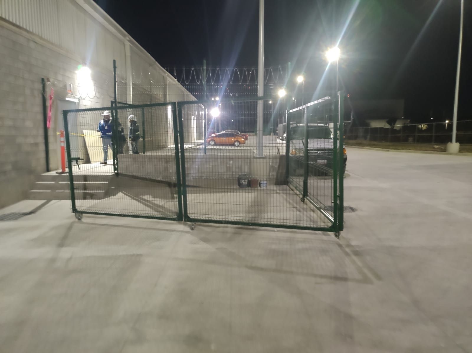 Escena nocturna: Zona verde vallada con puertas enrollables, edificio y trabajadores, un coche visible a lo lejos.
