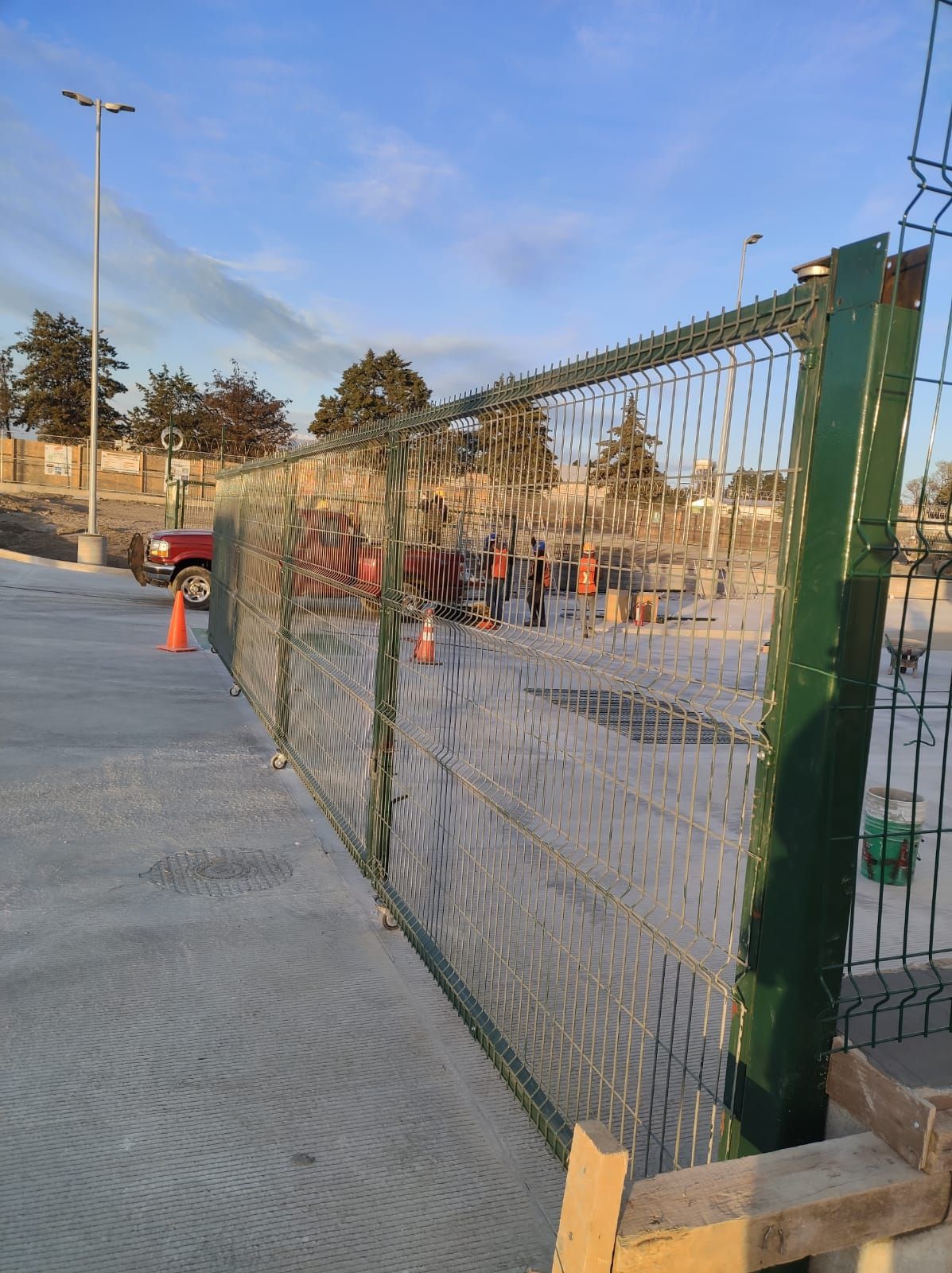 Una valla de alambre verde rodea una obra en construcción. Suelo de hormigón con trabajadores y equipo visibles.