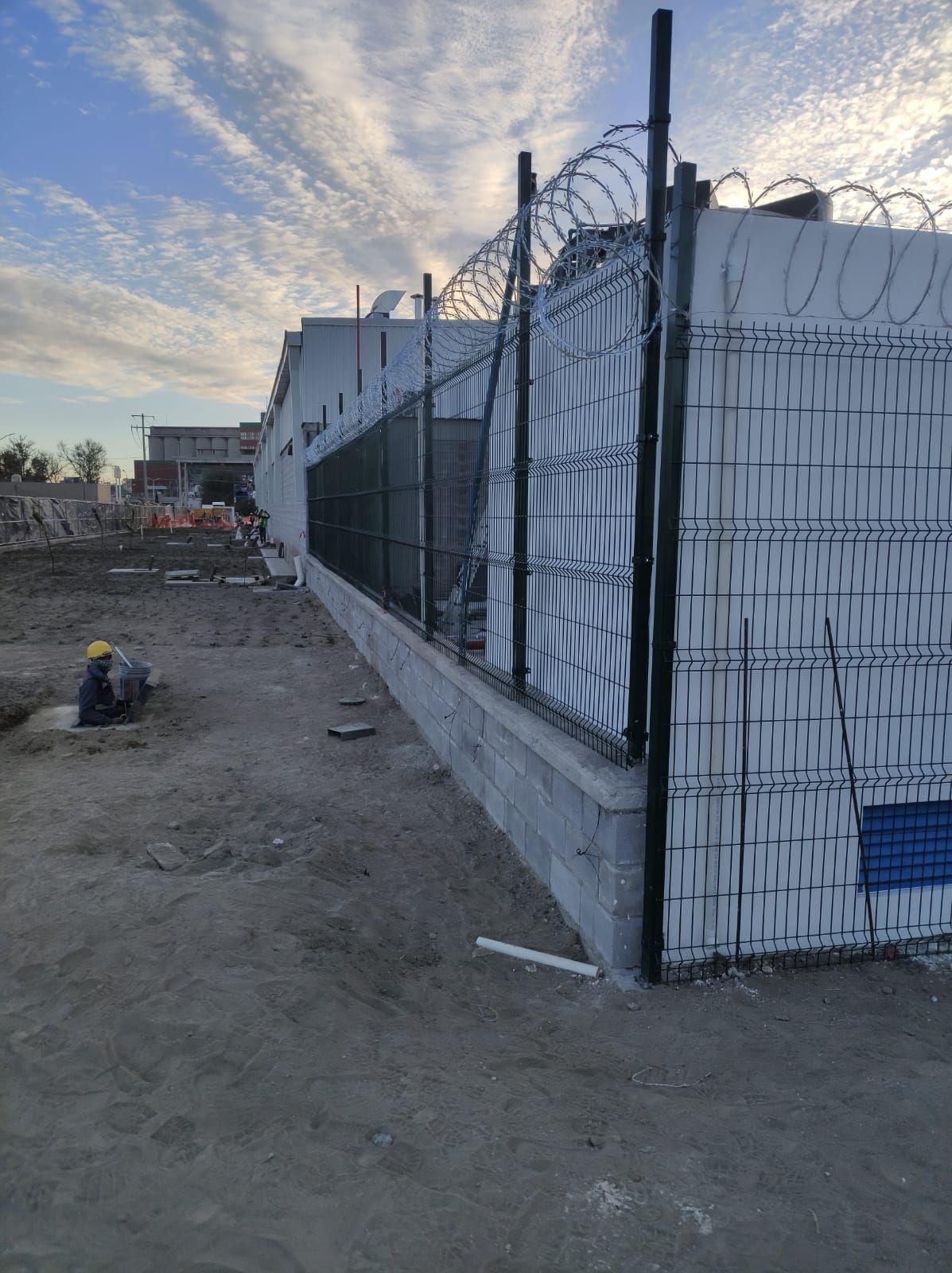 Sitio de construcción con valla de seguridad rematada con alambre de púas, un trabajador y cielo nublado.