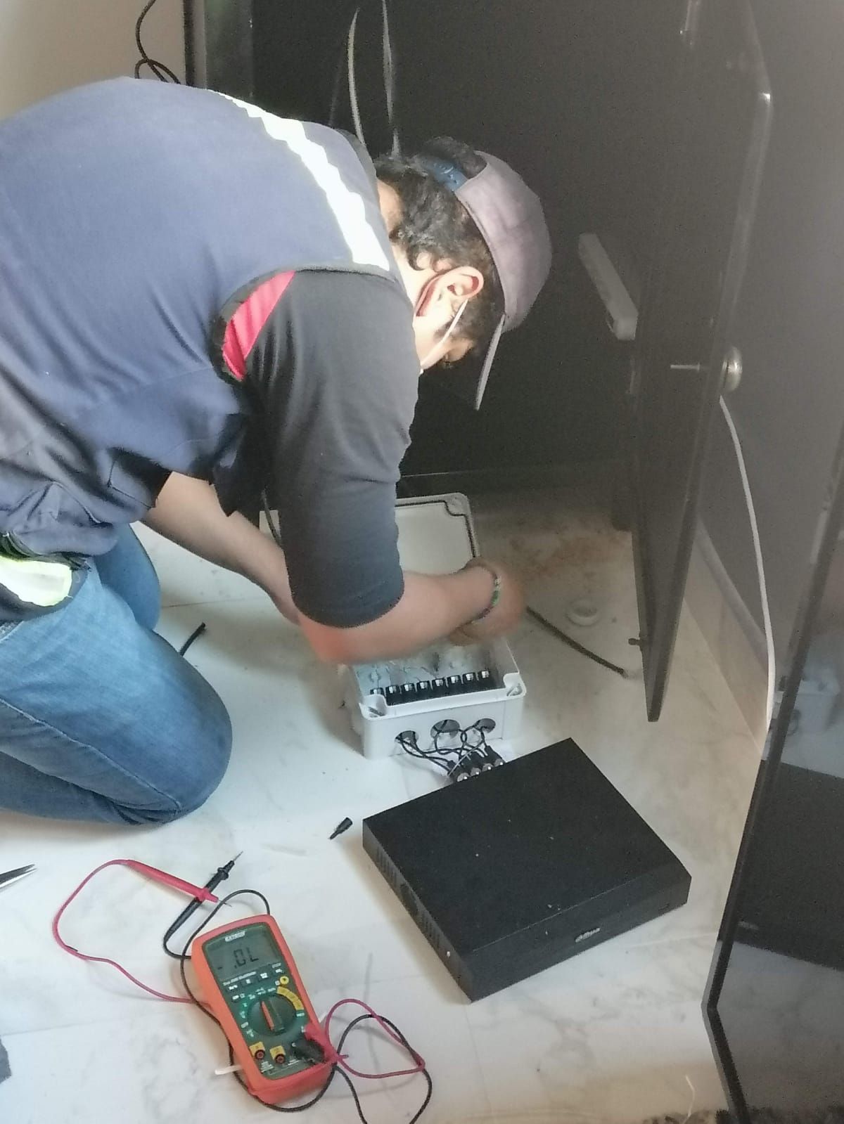 Un hombre con gorra trabaja en el cableado cerca de un dispositivo negro y un medidor en el suelo, cerca de un gabinete negro.