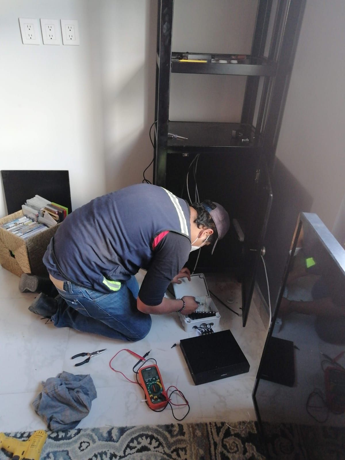 Hombre arrodillado, trabajando con electrónica frente a un rack de servidores negro. Cables, herramientas y equipos esparcidos por el suelo.