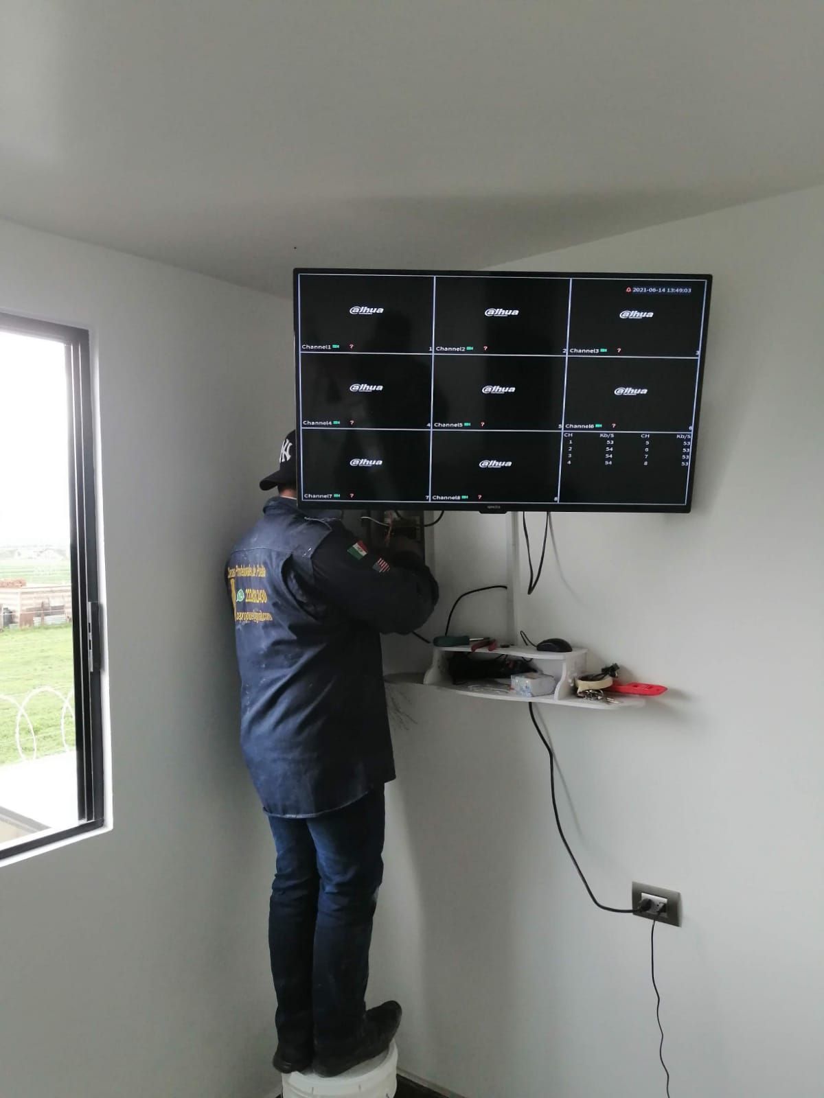 Una persona instalando un sistema de seguridad en una pared con una pantalla montada que muestra nueve imágenes de cámaras.