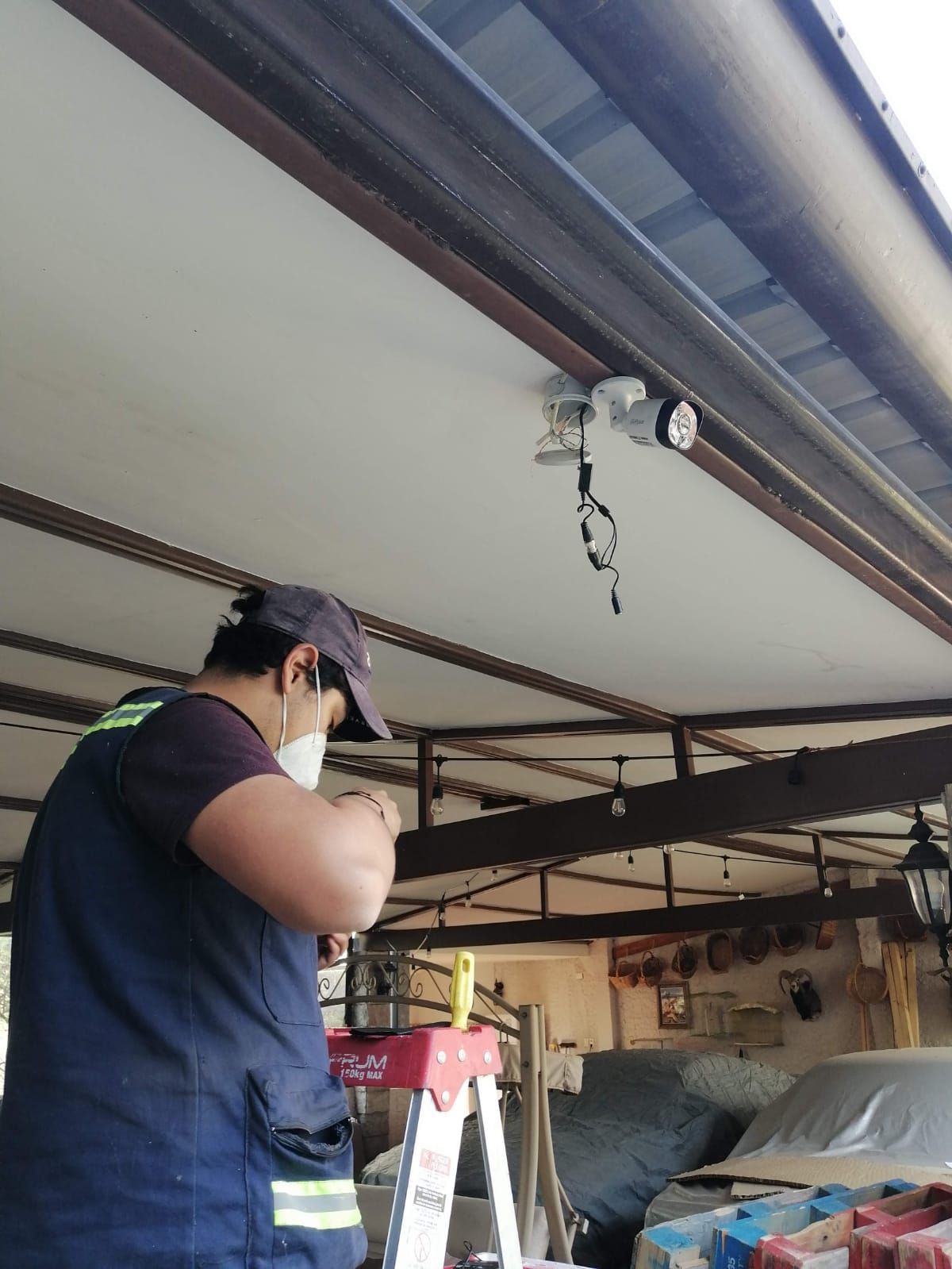 Hombre en una escalera instalando una cámara de seguridad bajo el techo de una cochera.