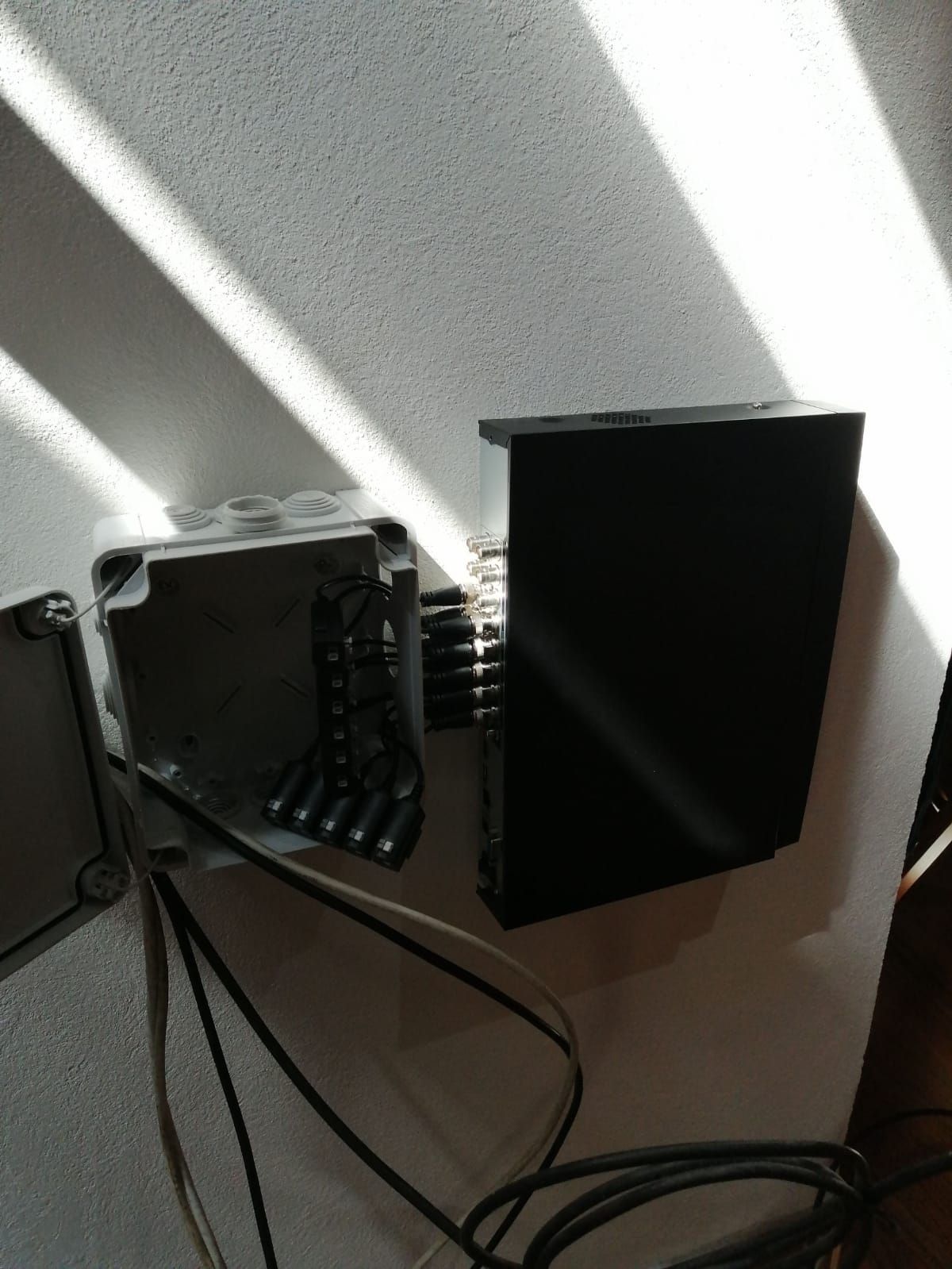 Una caja de conexiones eléctricas abierta conectada a un dispositivo negro en una pared blanca, con cables y luz solar.