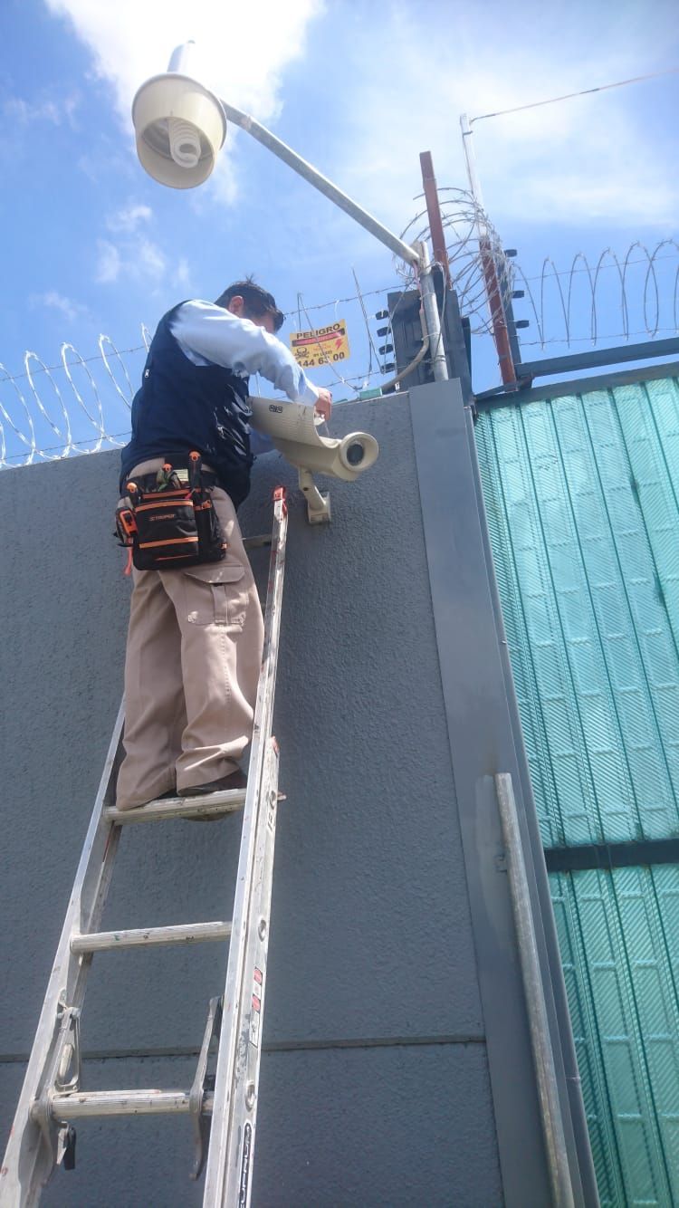 Persona en una escalera instalando una cámara de seguridad en un muro coronado con alambre de púas. Cielo azul.