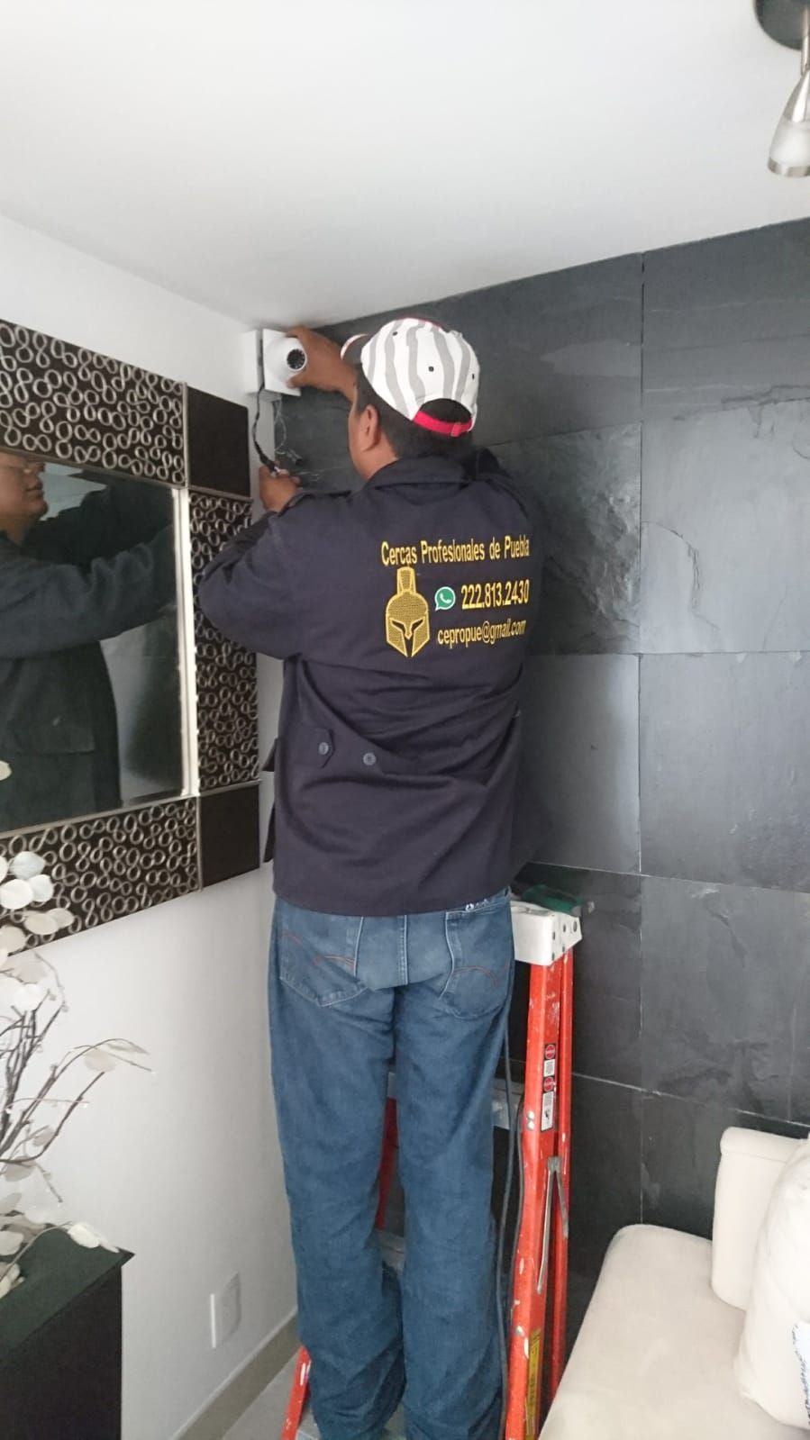 Hombre con jeans azules y chaqueta en una escalera, instalando algo en una pared.