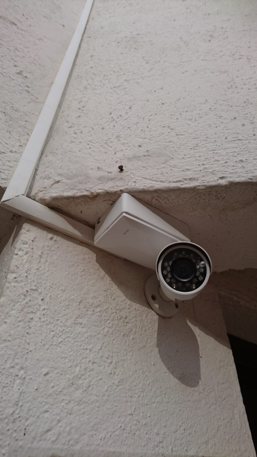 Cámara de seguridad blanca montada en una pared texturizada blanca, cerca de un conducto de cable blanco.