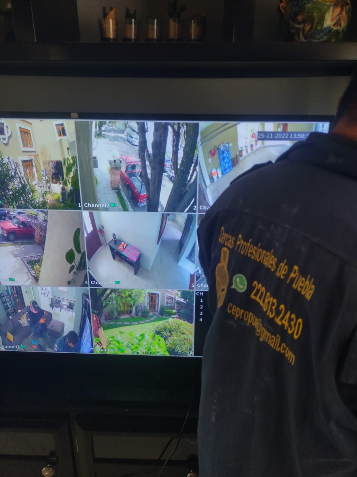 Un hombre con camisa negra monitorea las cámaras de seguridad de una casa. La pantalla muestra varias vistas de las cámaras: árboles, coches y exteriores de edificios.