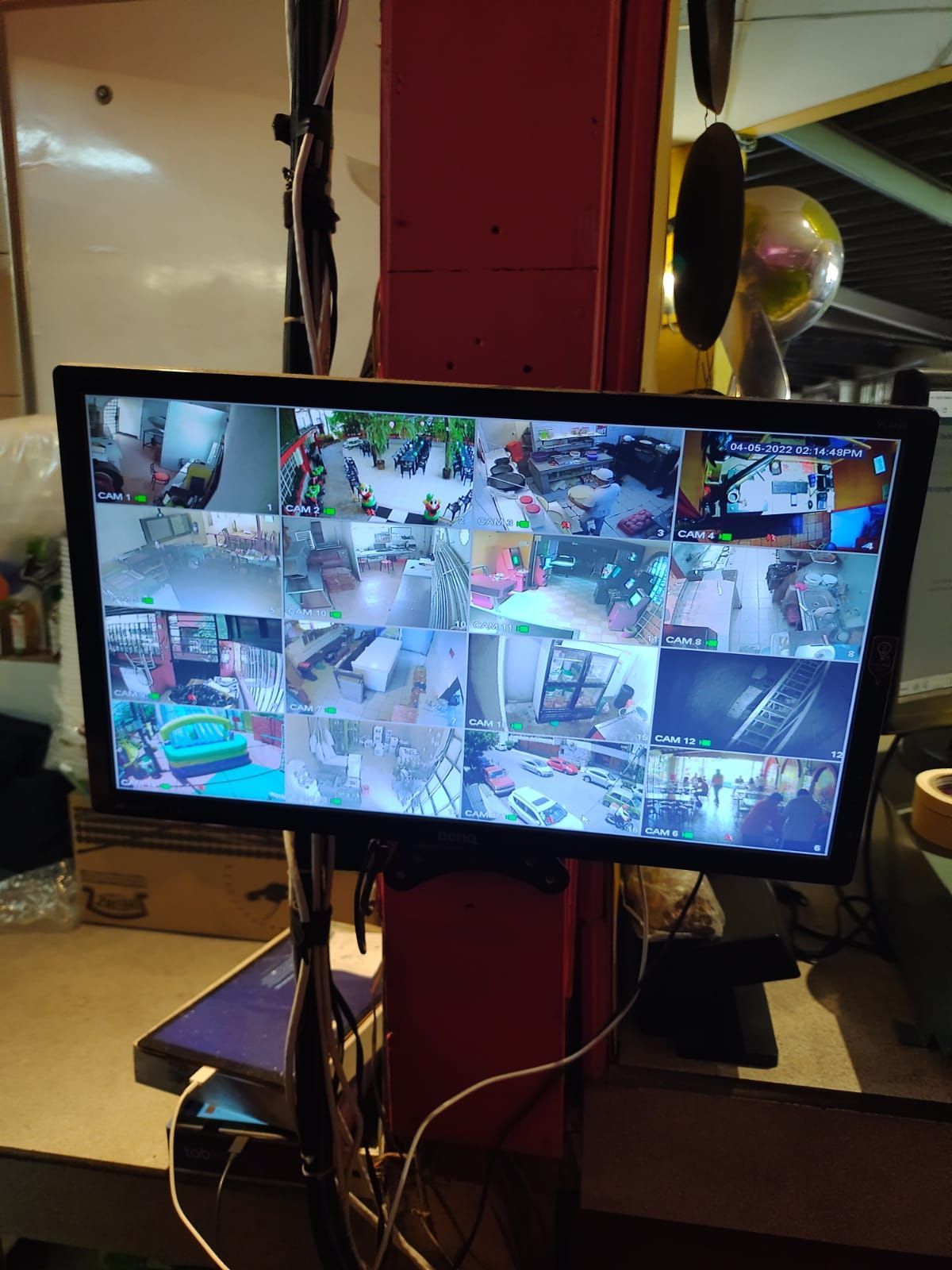 Monitor con múltiples vistas de cámaras de seguridad. Montado en un poste rojo, muestra áreas interiores y exteriores.
