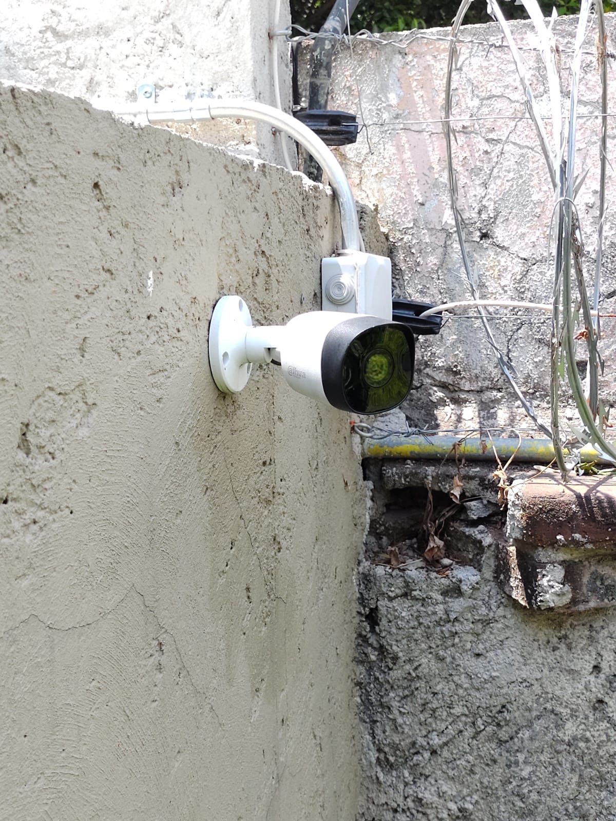 Cámara de seguridad blanca instalada en una pared clara con textura y cableado. La cámara está junto a una pared de ladrillos y alambre de púas.