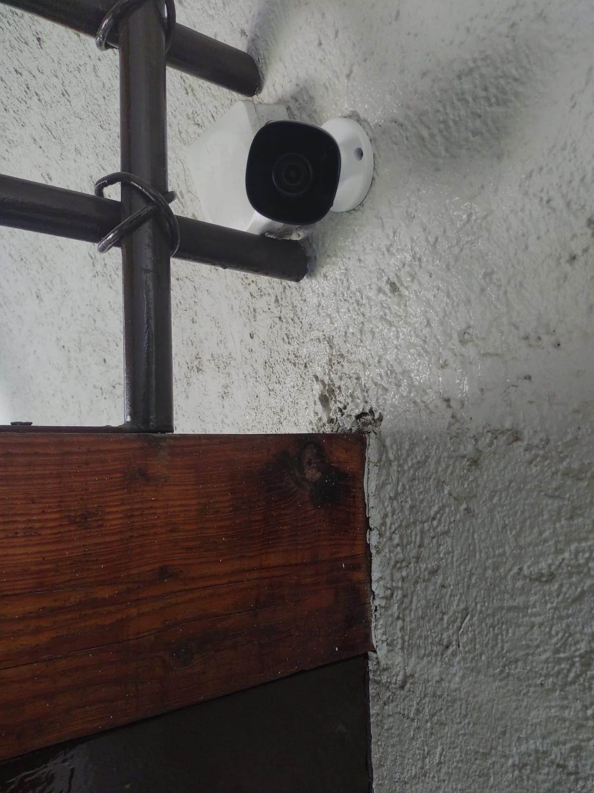 Cámara de seguridad montada en una pared gris texturizada cerca de una viga de madera y una barandilla de metal.