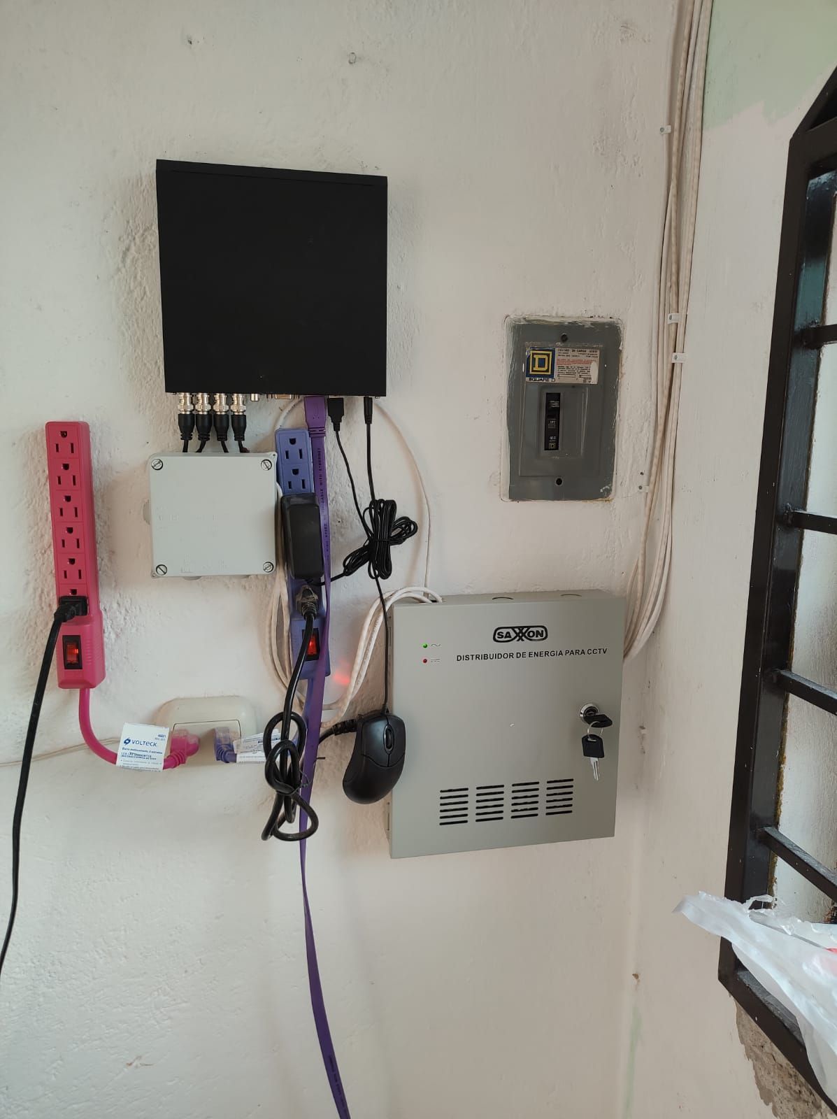Electrónica montada en la pared: una caja negra, cajas grises, una regleta de enchufes y un panel eléctrico en una pared blanca.