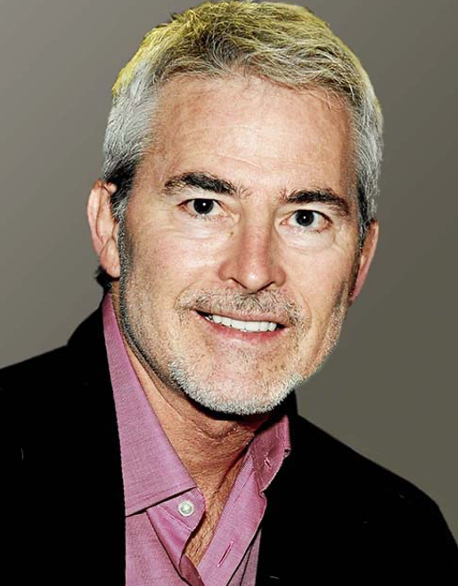 A man with gray hair and a beard is wearing a pink shirt and a black jacket.