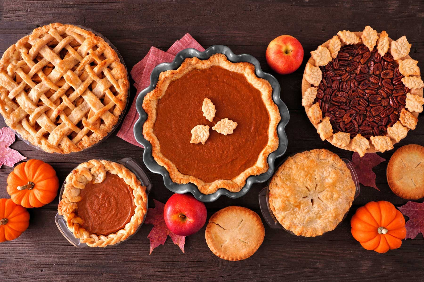 Assorted pies, pumpkins, and apples on a wooden table, fall harvest theme.