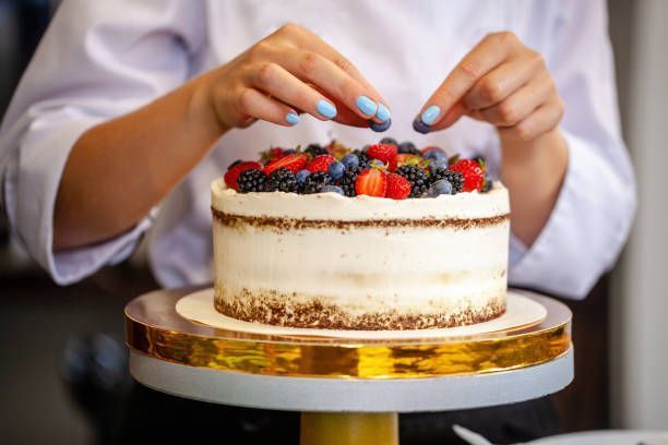 Baker decorates a layered cake with fresh berries on a golden cake stand.