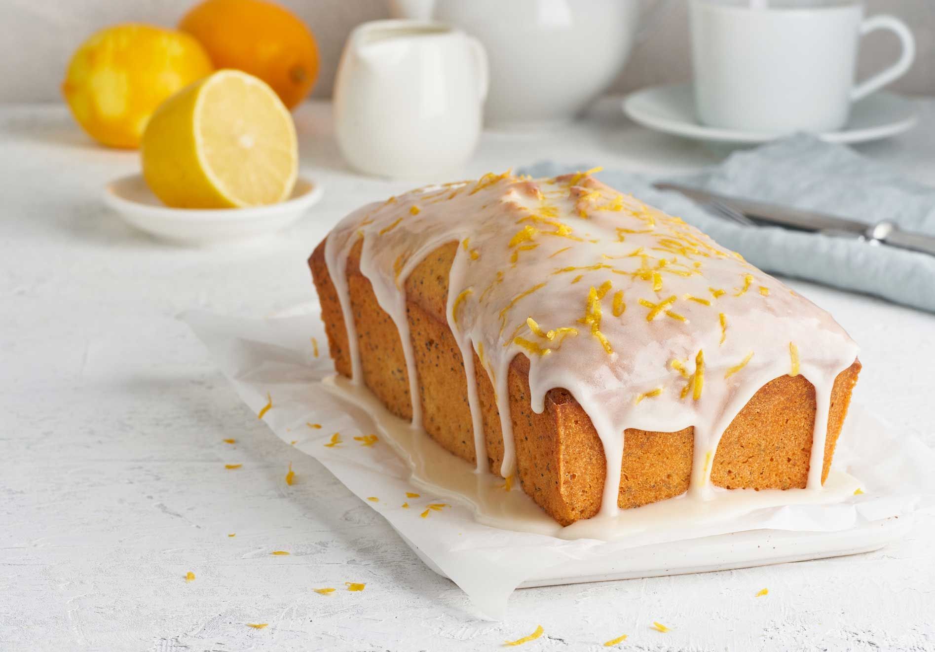 Lemon loaf cake with white glaze and lemon zest on a white surface, with lemons and a cup in the background.