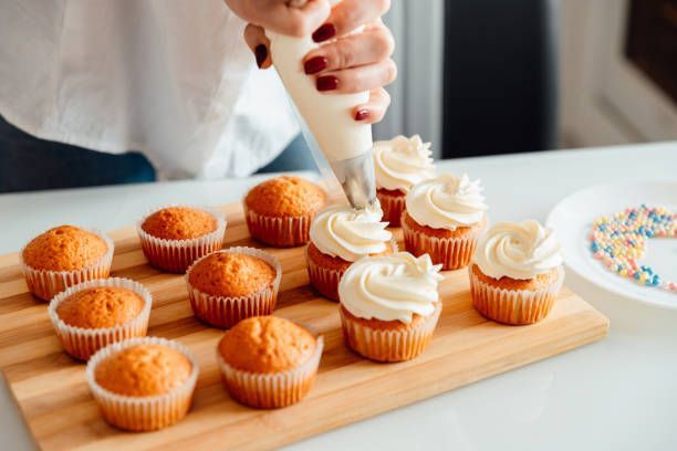 Person piping white frosting onto cupcakes on a wooden board.