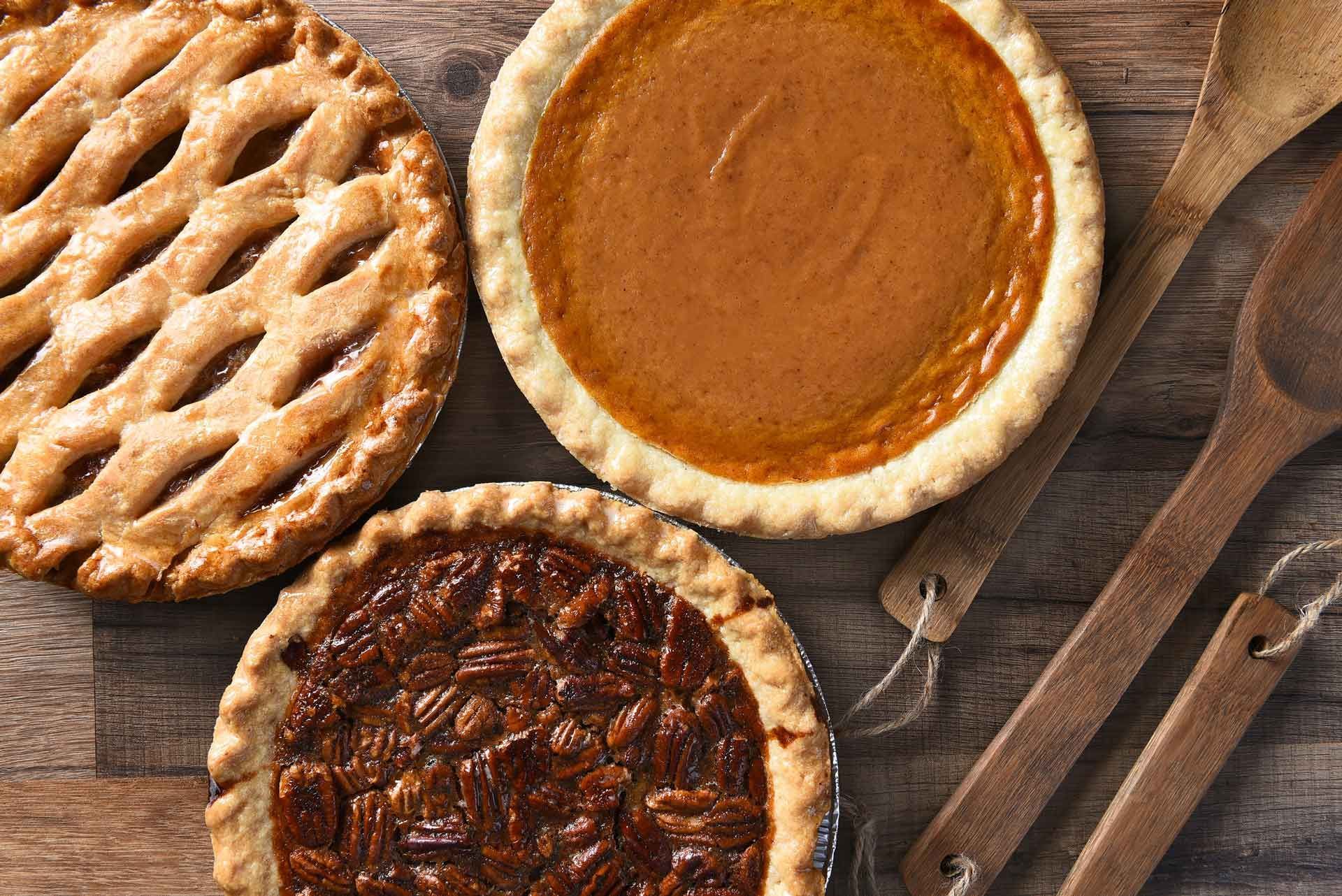 Three pies: apple lattice, pumpkin, pecan, with wooden spoons on a wood table.