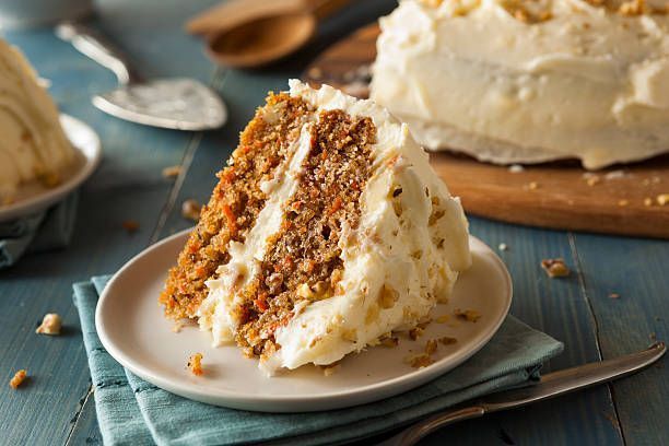 Slice of carrot cake with white frosting on a white plate, wooden board and a blue surface.