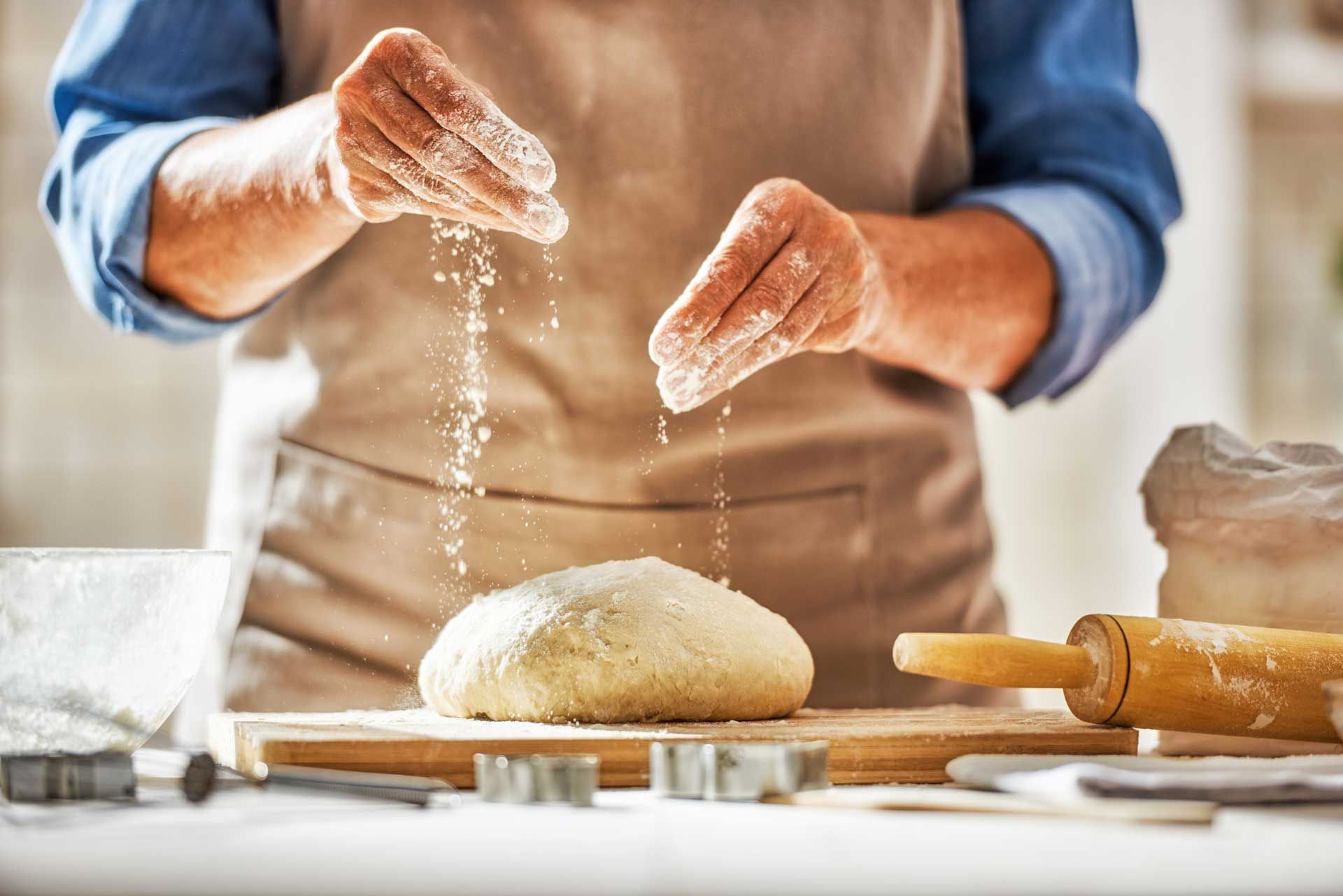Person in apron sprinkling flour onto dough. Rolling pin and cookie cutters on the counter.